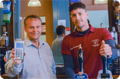 Owner of a pub holding up card payment machine with member of staff pulling a pint